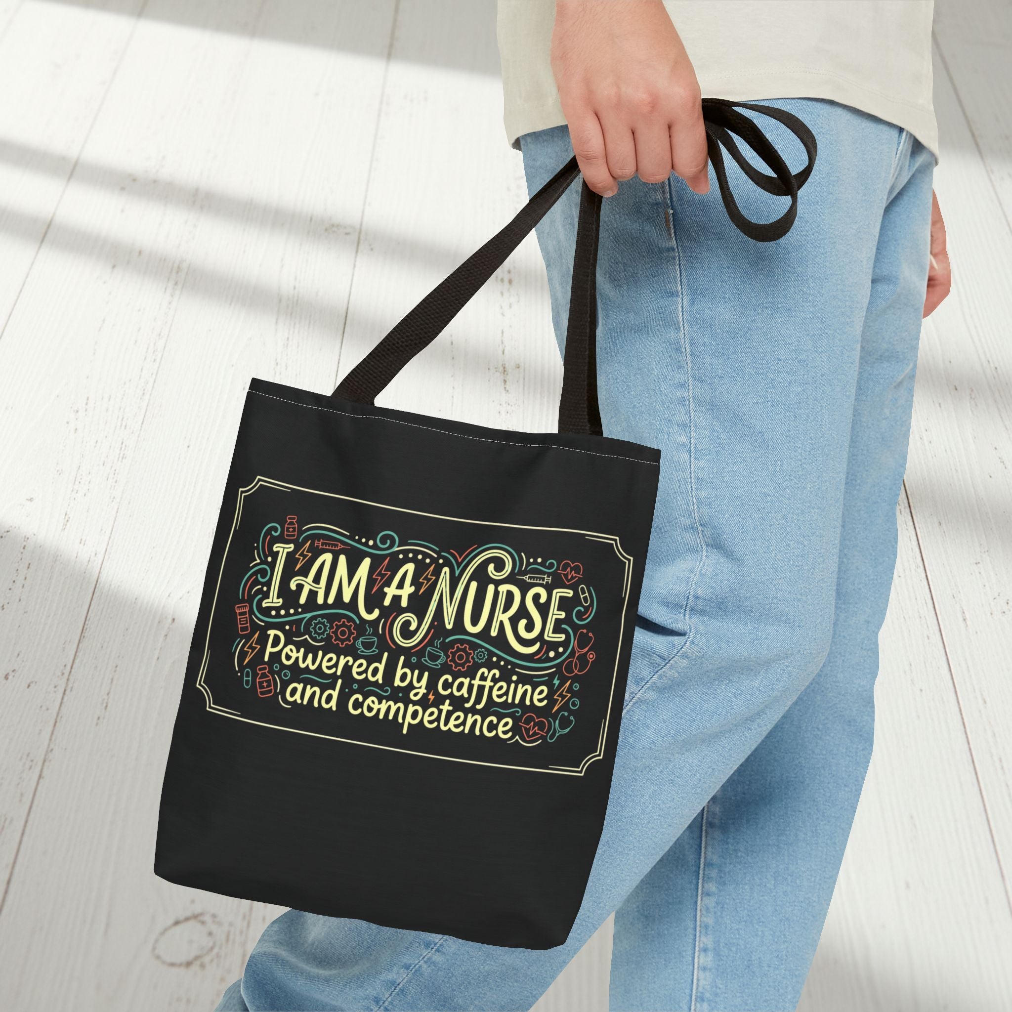 Black tote bag with 'I AM A NURSE, powered by caffeine and competence' text held by a person wearing blue jeans.